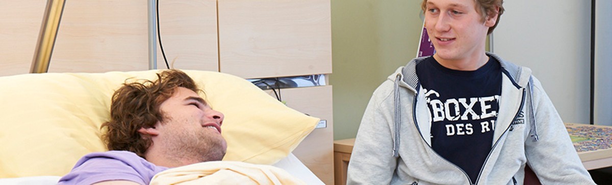 Ein im Bett liegender Patient unterhält sich mit seinem Besuch, der neben ihm sitzt.
