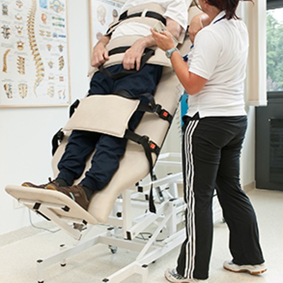 Ein Patient steht am Stehbett etwa 45° mit einer Therapeutin zu ihm gewandt an seiner Linken