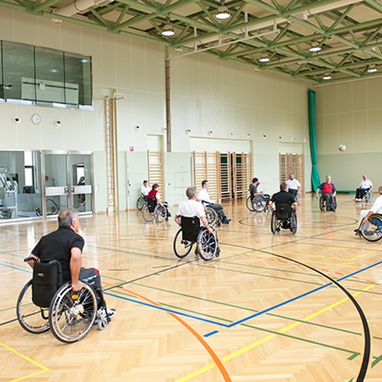 Rollstuhltraining: Eine Gruppe von Rollstuhlfahrern steht gleichmäßig verteilt in der Turnhalle. Ein fliegender Volleyball ist zu sehen.