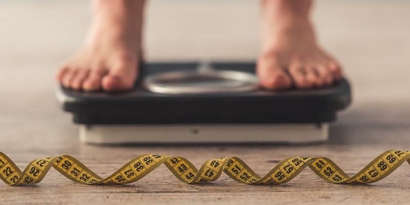 Füße auf Waage und davor liegt ein ausgerolltes Maßband; Foto: VGstockstudio, Shutterstock.com