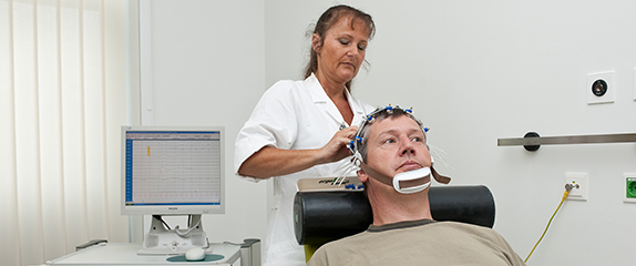 Eine  Fachkraft  positioniert EEG-Elektroden am Kopf eines auf dem Untersuchungsstuhl liegenden Patienten