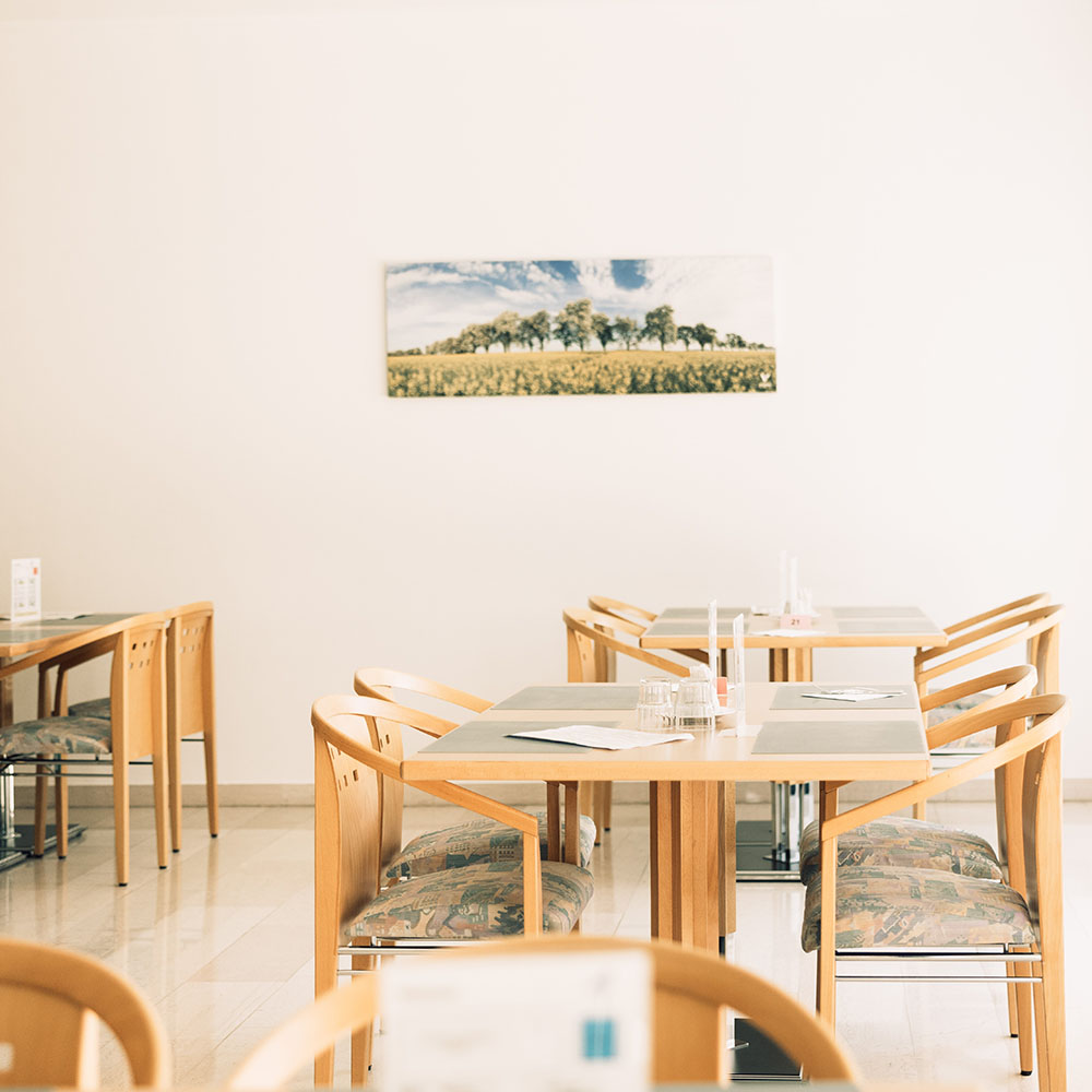 Schlicht gehaltener Speisesaal des Gesundheitszentrums: Zu sehen sind zwei kleinere Tische mit je vier gepolsterten Holzsesseln; Der Boden ist hell gefliest, an der ebenso hell gestrichenen Mauer hängt ein Landschaftsbild.