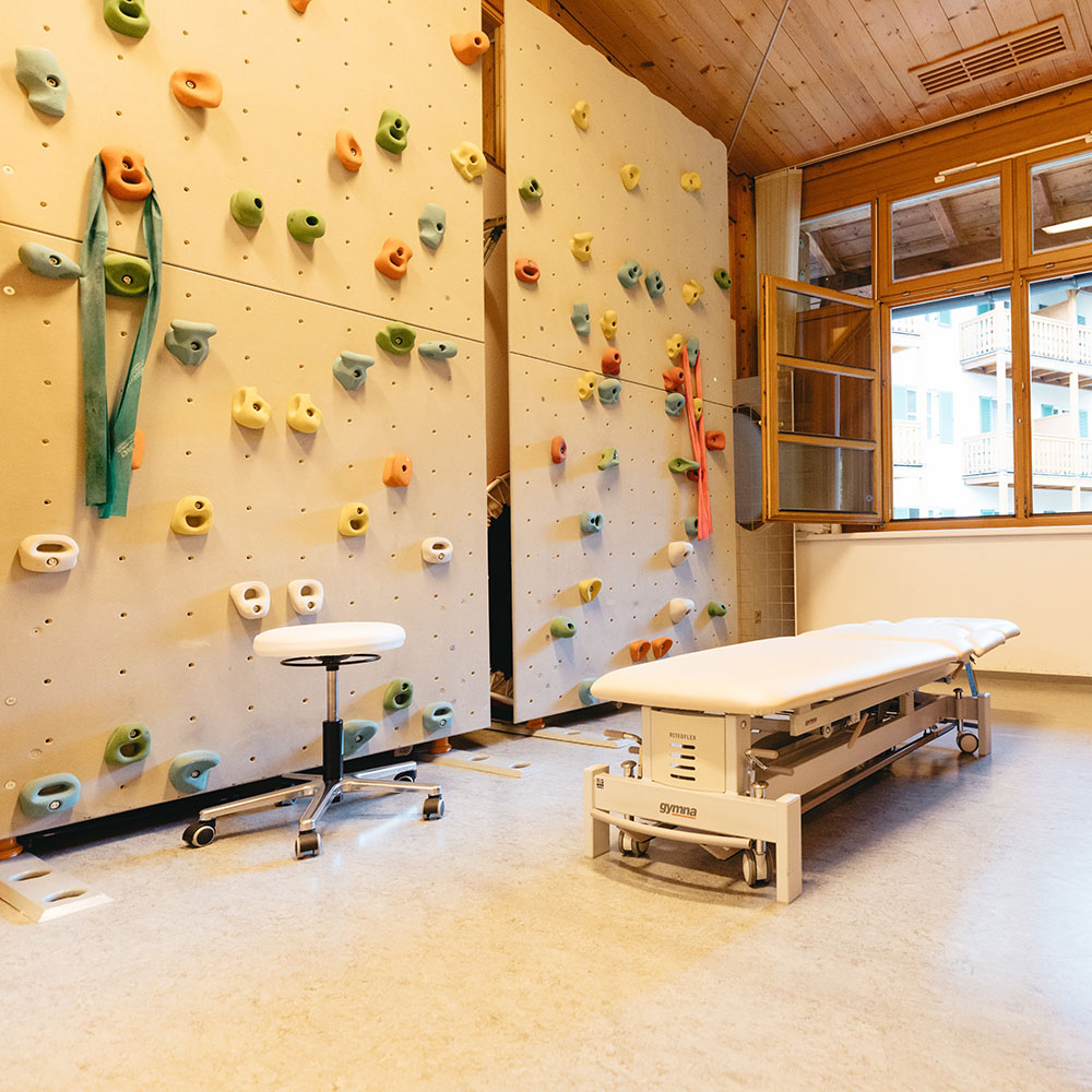 Großer Therapieraum, linkerhand mit therapeutischer Kletterwand, heller Boden, Holzdecke. Eine weiße Behandlungsliege mit Hocker steht vor einem geöffneten Fenster mit Blick auf eine Gebäudefront.